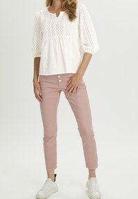 Donna che indossa una blusa bianca a maniche a sbuffo in pizzo, pantaloni slim a vita alta rosa pallido con bottoni decorativi e sneakers bianche su sfondo bianco.