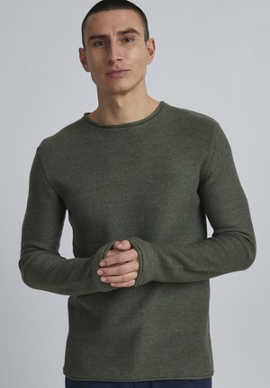 Grønn teksturert strikkegenser med lange ermer, rund hals og tommelholdere ved mansjettene, vist brukt av en modell med kort hår.