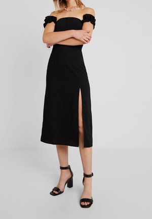 Mujer con vestido midi negro con hombros descubiertos y abertura frontal, combinado con sandalias negras de tacón alto y tiras en el tobillo.