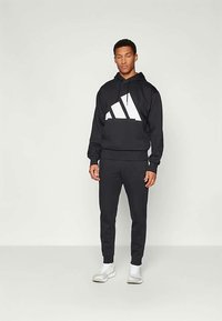 Čierna mikina s kapucňou a bielym trojuholníkovým logom, v kombinácii s čiernymi športovými nohavicami. Zladené s bielymi športovými topánkami. Hladká textúra látky.