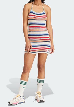 Femme portant une robe dos-nu rayée multicolore, des chaussettes blanches montantes aux genoux avec des rayures vertes, et des chaussures de sport blanches avec des accents colorés.