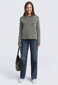 Sivo dolgoročni pulover z enakomernimi črtami in rebrasto teksturo, kombiniran z modrimi kavbojkami z ravnimi hlačnicami in belimi supergami. Drži črno torbico čez ramo.