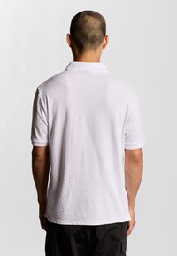 Vit kortärmad polo med en texturerad, bikakestruktur. Har en klassisk krage och ribbade ärmslut. Visas från baksidan.