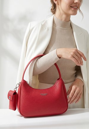 Donna con top beige e blazer bianco che tiene in mano una borsa rossa Diana & Co Firenze con una piccola pochette attaccata su una superficie bianca.