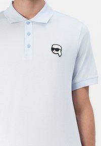 Lyseblå poloskjorte laget av myk stoff, med en tre-knapps placket og et svart-hvitt grafikkmerke av et ansikt med solbriller.