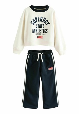 Witte cropped sweatshirt met lange mouwen met tekst "Superdry State Athletics" en vlag, gecombineerd met marineblauwe broek met trekkoord, witte strepen en vlagpatch.