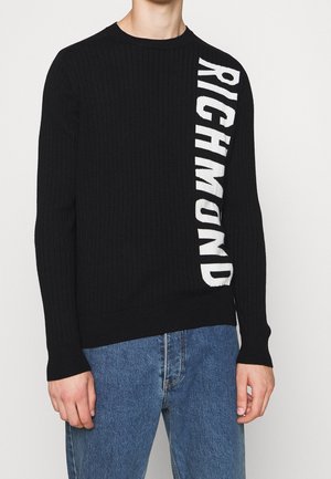 Pull côtelé noir avec "RICHMOND" en grandes lettres blanches verticales sur le côté droit, porté avec un jean bleu.