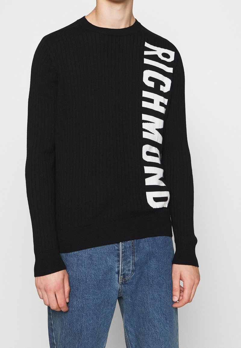 Pull côtelé noir avec "RICHMOND" en grandes lettres blanches verticales sur le côté droit, porté avec un jean bleu.