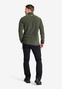 Fleece-tröja i mörkgrön med hög krage. Slim-fit design, långa ärmar och subtil logotypdetalj på ryggen. Svarta byxor och skor.