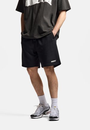 Mężczyzna noszący czarne szorty z logo "PROSTO", ciemną, oversize koszulkę, białe skarpetki oraz białe i czarne sneakersy w wzory, stojący z rękami w kieszeniach.