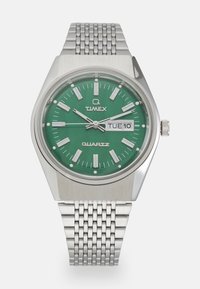 Reloj de cuarzo Timex con un dial verde, marcadores de hora en tono plateado y una pantalla de fecha. Presenta una pulsera de malla de acero inoxidable y una caja hexagonal.