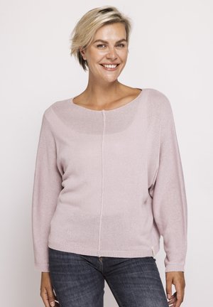 Smiling woman with short blonde hair wearing a light pink knit sweater and dark blue jeans standing against a plain background.