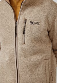 Beige Zip-Up-Pullover mit strukturiertem Strick, Brustreißverschlusstasche und „s.O Elevated Casual Est. 1969“-Aufnäher auf der linken Brust.