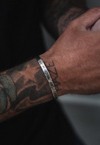 Hõbedane mansettbracelet, millel on löökpind ja graveeritud märgised, kantud tätoveeritud randmel, millel on nähtavad nahatooni variatsioonid.
