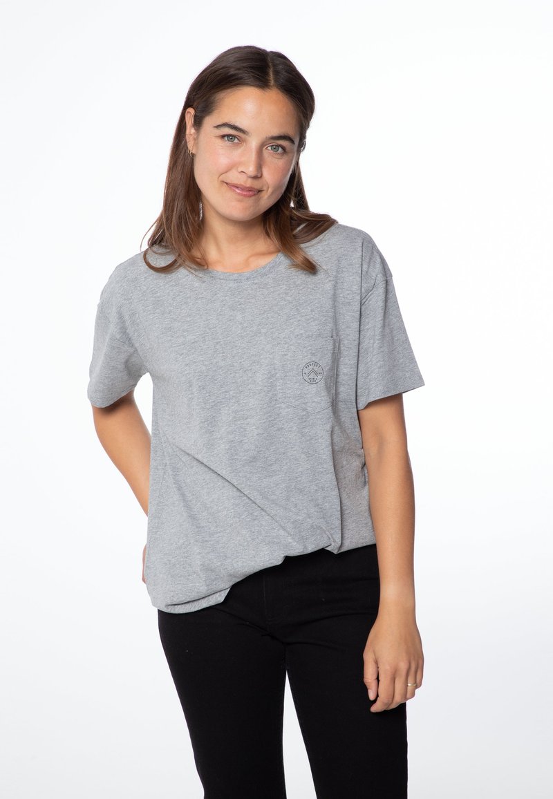 Camiseta de algodón gris con un corte relajado, cuello redondo, mangas cortas y un pequeño logo en el lado izquierdo del pecho. Combinada con pantalones negros.