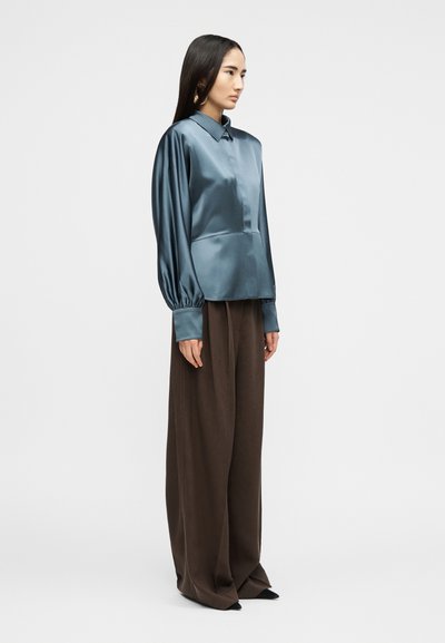 Femme debout portant un chemisier en satin bleu brillant avec des manches bouffantes et des poignets larges, associé à un pantalon large et ample marron.