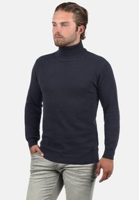 Navy coltrui in een gemêleerd breipatroon, met geribbelde manchetten en zoom, gecombineerd met lichtgrijze jeans.