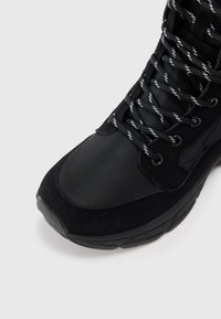 Black high-top sneaker with black and white patterned laces on a light background, showing toe and part of the sole.