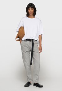 Camiseta blanca de gran tamaño, pantalones vaqueros grises con cinturón negro y zapatos negros tipo slip-on. Bolsa de hombro marrón con detalle de cierre. Fondo liso.