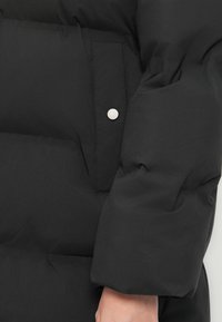 Nahaufnahme eines schwarzen gepolsterten Jackenärmel und einer Tasche mit silbernem Knopf, die Nähdetails zeigt und eine teilweise sichtbare Hand.