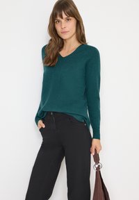 Suéter azul verdoso con cuello en V, de mangas largas, tejido texturizado y corte relajado. Combinado con pantalones negros y sosteniendo un bolso marrón con acentos metálicos.