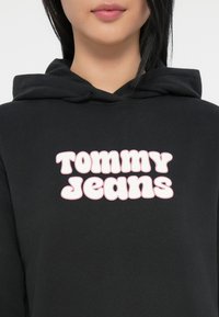 Sweatshirt preta com um grande logótipo em texto branco arredondado que diz "TOMMY JEANS" com detalhes em rosa. Tecido macio com um corte solto.