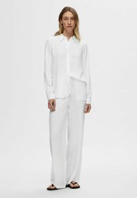 Witte, lang-sleeve button-up shirt gecombineerd met losse witte broek. Zacht, lichtgewicht stof met een subtiele textuur. Beschikt ook over een trekkoord in de taille.