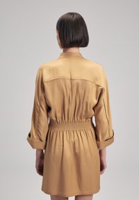 Vestido caqui con cintura ceñida, mangas largas y cuello con solapa. Tejido suave con un ligero brillo y puños doblados.