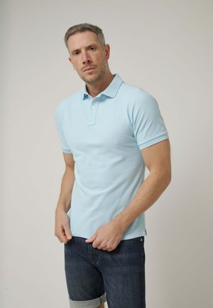 Hombre con cabello corto, vestido con una camisa tipo polo azul claro y vaqueros oscuros remangados, de pie frente a un fondo beige liso.