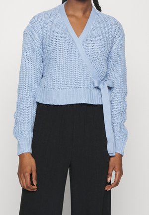 Personne portant un pull cache-cœur tricoté bleu clair noué sur le côté et un pantalon noir ample, debout devant un fond uni.