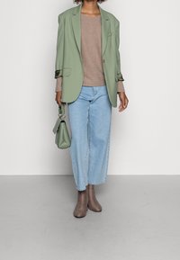 Person trägt ein salbeigrünes Blazer über einem taupefarbenen Pullover, hellblaue weite Jeans, braune Ankle Boots und hält eine passende grüne Handtasche.