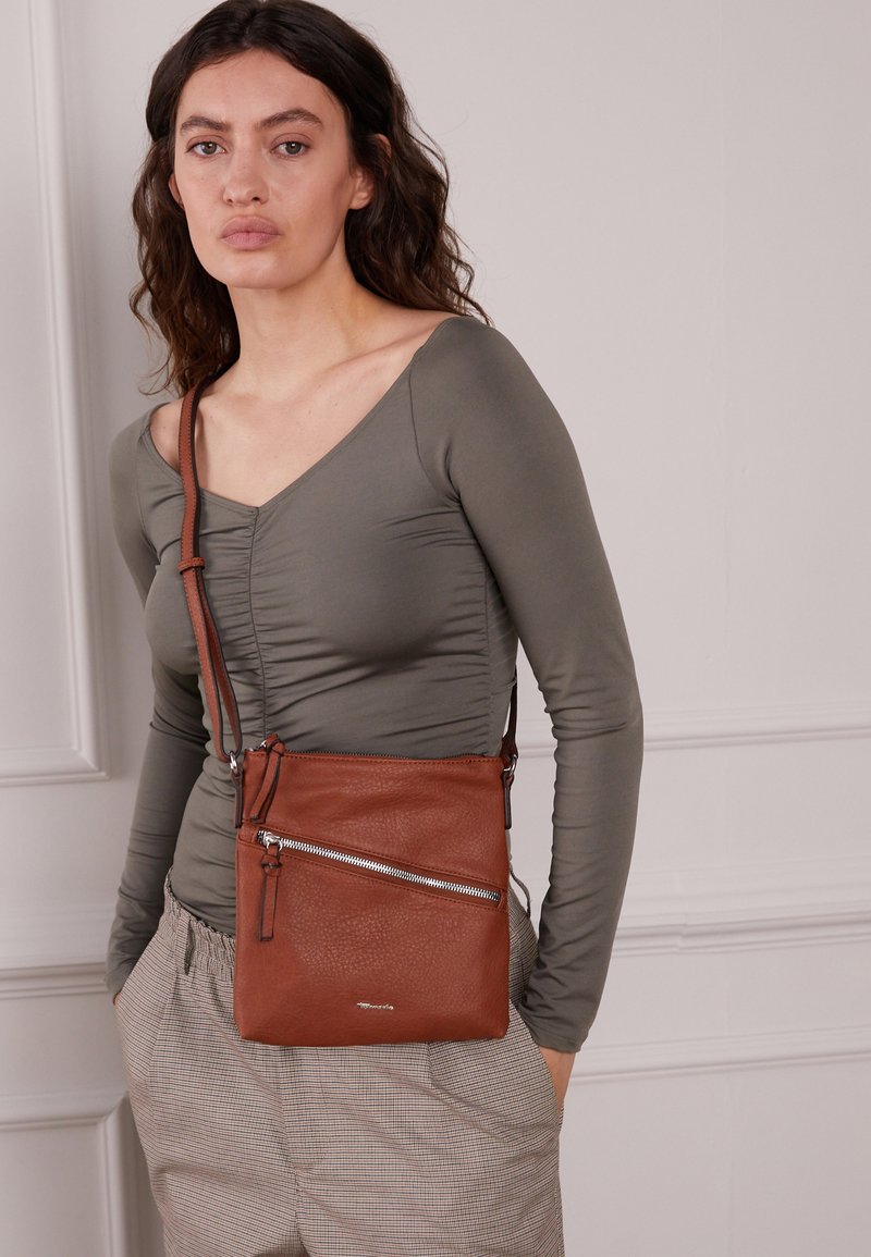 Braune Leder-Crossbody-Tasche mit strukturierter Oberfläche, einer vorderen Reißverschlusstasche und einem verstellbaren Tragegurt. Getragen mit einem figurbetonten olivgrünen Oberteil.