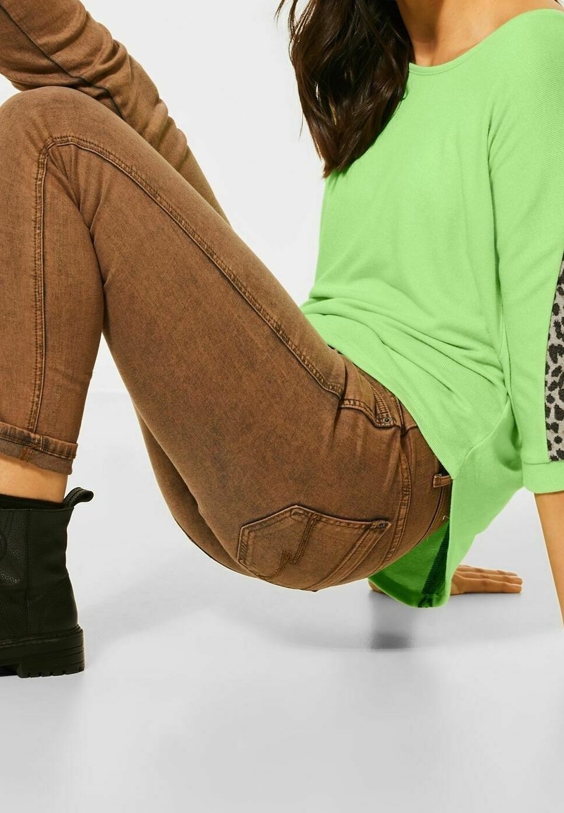 Bruna denimbyxor med smal passform, kombinerade med en ljusgrön skjorta. Skjortan har svarta leopardmönstrade detaljer på ärmarna. Svarta ankelstövlar bärs.