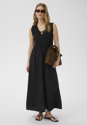 Femme portant une robe maxi noire sans manches, des sandales noires, des lunettes de soleil foncées, tenant un grand sac en daim marron sous un bras.