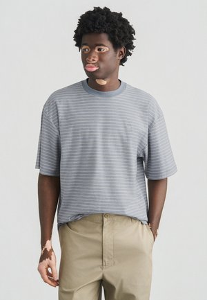 Szary t-shirt w paski z krótkim rękawem i okrągłym dekoltem, wykonany z miękkiej tkaniny. Połączony z jasnobeżowymi spodniami o luźnym kroju.