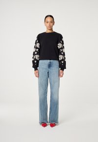 Zwarte sweatshirt met geborduurde witte bloemen- en sterontwerpen op de mouwen, gecombineerd met lichtblauwe wijde jeans en rode puntige schoenen.