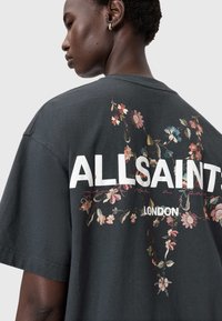 Harmaa puuvillainen t-paita, jossa on kukkakuvio pinkin, sinisen ja vihreän sävyissä, ja valkoinen teksti "ALLSAINTS" ja "LONDON" paidan takana.