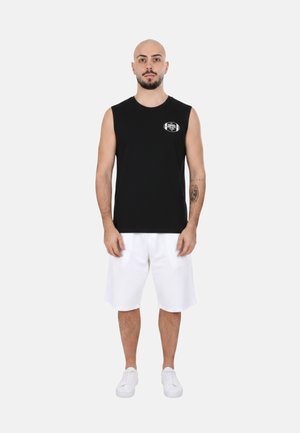 Uomo in piedi, indossa una maglietta nera senza maniche con un piccolo logo bianco, pantaloncini bianchi e sneakers bianche, su uno sfondo chiaro e uniforme.