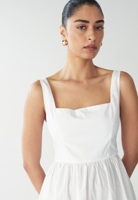 Vestido de algodón blanco con escote cuadrado, cuerpo ajustado y falda acampanada. La modelo tiene el cabello recogido y lleva pendientes de aro dorados.