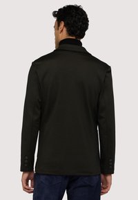 Donkergroene jas van geweven stof, met een nietched kraag en drie knopen op de mouwen. Gladde afwerking en op maat gemaakte pasvorm.