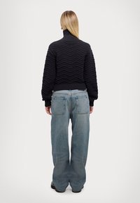Pull noir en maille avec un motif zigzag texturé, col montant et épaules tombantes ; associé à un jean denim bleu clair à jambes larges.
