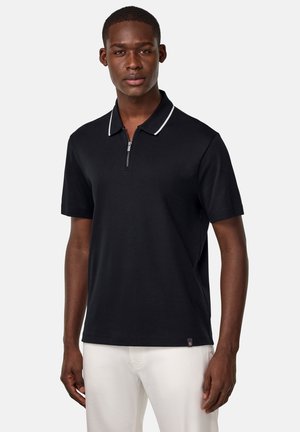 Polo negro de manga corta con cuello con cremallera, presenta detalles en blanco a lo largo del cuello, fabricado en una tela suave, exhibido en un modelo frente a un fondo liso.