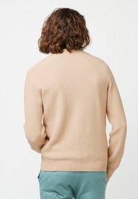 Beige gestrickter Pullover mit einem strukturierten Muster und gerippten Bündchen, von hinten betrachtet. Der Pullover wird mit hellblauen Hosen kombiniert.