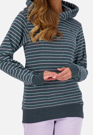 Femme portant un sweat à capuche à rayures bleues avec des trous pour les pouces, mains jointes devant, et pantalon violet clair, vue du cou jusqu’aux hanches.