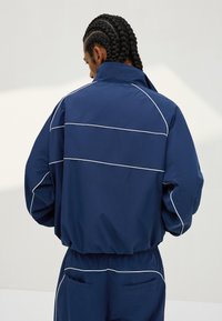 Oseba s pletenicami, oblečena v mornarsko modro trenirko z belim obrobjem, obrnjena stran, na svetlem, enobarvnem ozadju.