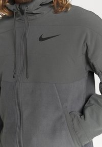Giacca Nike grigia con chiusura a zip, texture in felpa nella parte inferiore e logo ricamato sul petto. Cappuccio con cordini regolabili.