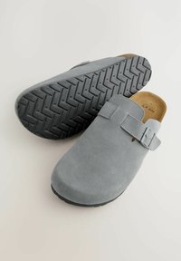 Grey suede clogs with a rounded toe, adjustable strap, and textured black rubber sole featuring a zigzag pattern for grip.