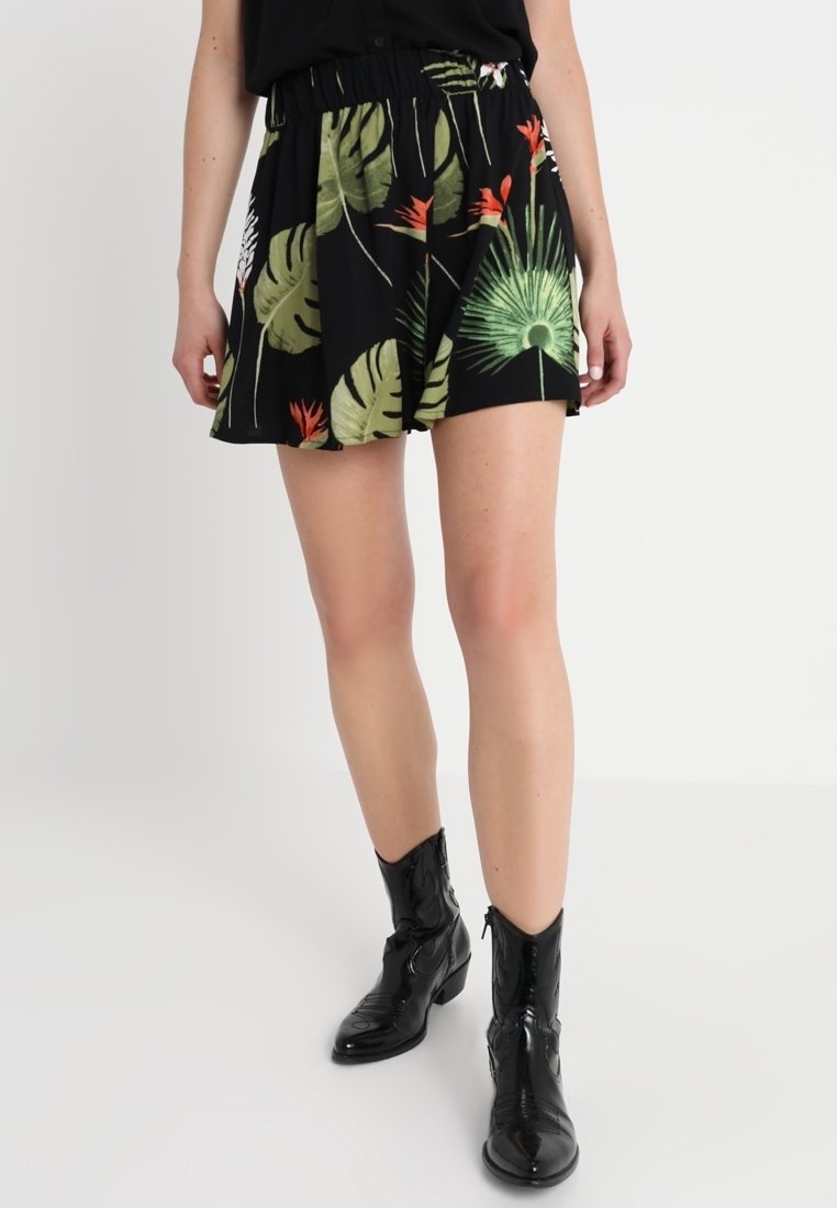 Femme portant un short noir à motifs de feuilles tropicales vertes et orange, assorti à des bottines noires brillantes, sur un fond blanc uni.