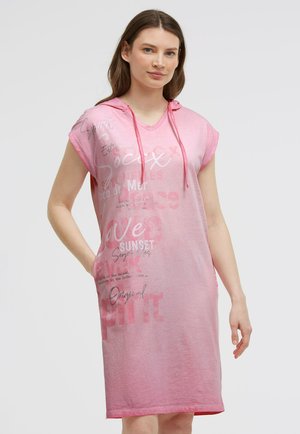 Young woman wearing a loose pink sleeveless hooded dress with printed text, standing with one hand in pocket against a plain background.