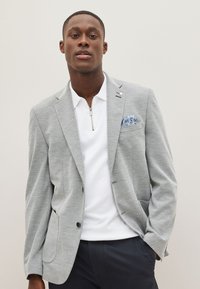 Blazer gris en tissu texturé, fermeture à un bouton et revers crantés. Polo blanc avec zip, pantalon bleu marine, et pochette bleue à motif.